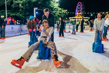 Couple ice skating. Girl being pushed on a kanga skating aid and throwing up a peace sign.