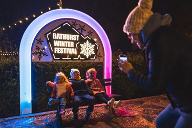 Three kids sitting in front of the Bathurst Winter Festival photo op sign and having their picture taken.