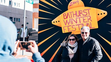 Couple having the picture taken in front of the 2019 Bathurst Winter Festival photo wall which is a painting of a bright yellow UFO with the words 2019 Bathurst Winter Festival written on it.