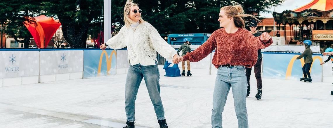 Two girls ice skating on the McDonald's Bathurst Ice Rink holding hands and dressed in warm knit jumpers.