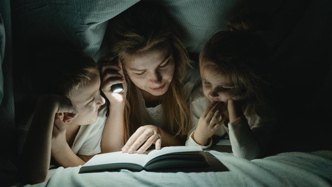 Three kids reading a book with a torchlight under the blankets of their bed.